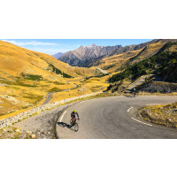 Cyclist on the corner of a road that winds through alpine grassland with mountain peaks in the distance on Marmot Tours full support road cycling tour