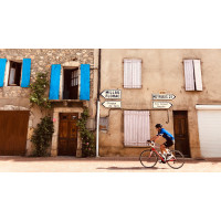 A cyclist in a blue jersey rides down a village road in rural france past a house with blue shutters on Marmot Tours supported road cycling holiday
